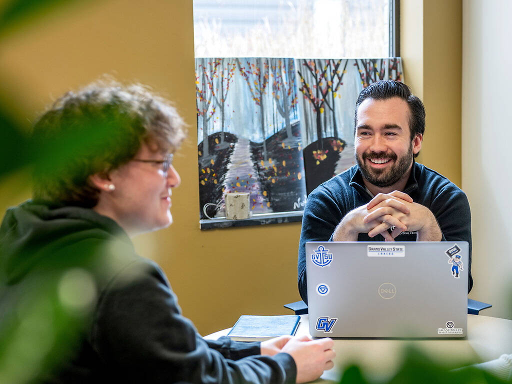 Two individuals engage in a lively conversation around a table, with one person smiling broadly while seated behind a laptop adorned with stickers.